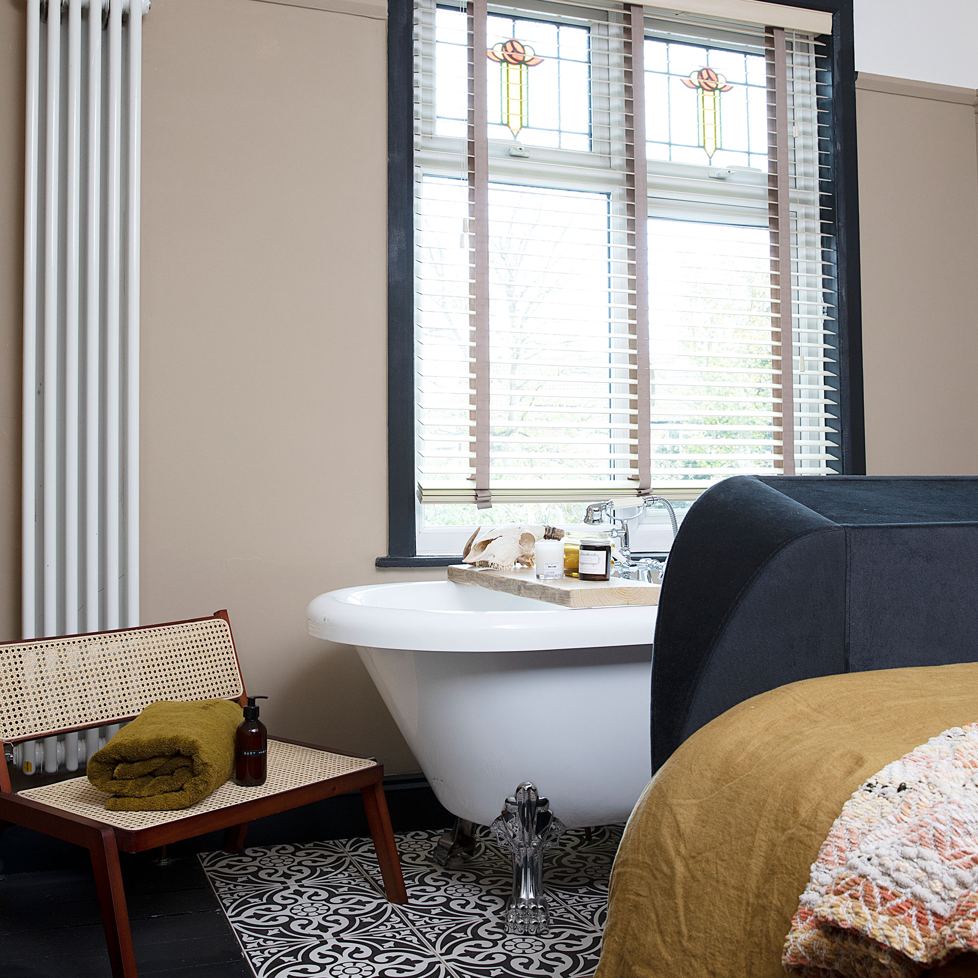 Kamar tidur netral dengan bak mandi berdiri bebas, jendela kaca patri, tempat tidur berlapis biru, lantai keramik